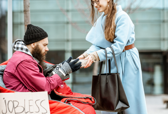 A photo of a woman in smart work clothes handing food to a man sitting on the street in a sleeping bag. wearing a beanie. His cardboard sign says jobless.