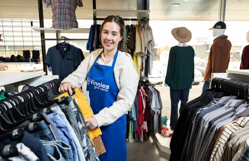 A photo of a Vinnies WA volunteer wearing a blue Vinnies apron while sorting through a clothing rack. She is smiling and looking at the camera. 