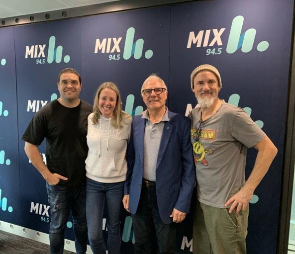 A group photo of the Vinnies WA State President together with 3 team members of Mix94.5 in front of a promo media wall displaying the logo for the radio channel. All four of them is smiling and facing the camera.