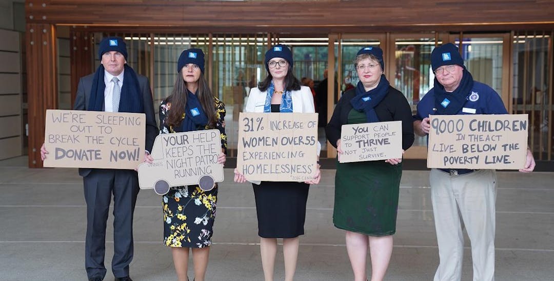 A photo of 5 people standing in a row, facing the camera and holding signs on cardboard about homelessness. They are wearing suits with beanies and scarves.