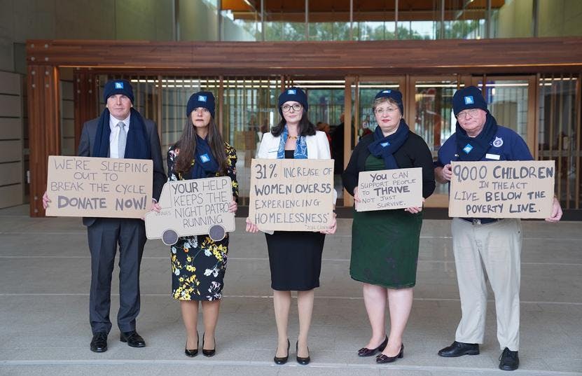 A photo of 5 people standing in a row, facing the camera and holding signs on cardboard about homelessness. They are wearing suits with beanies and scarves.