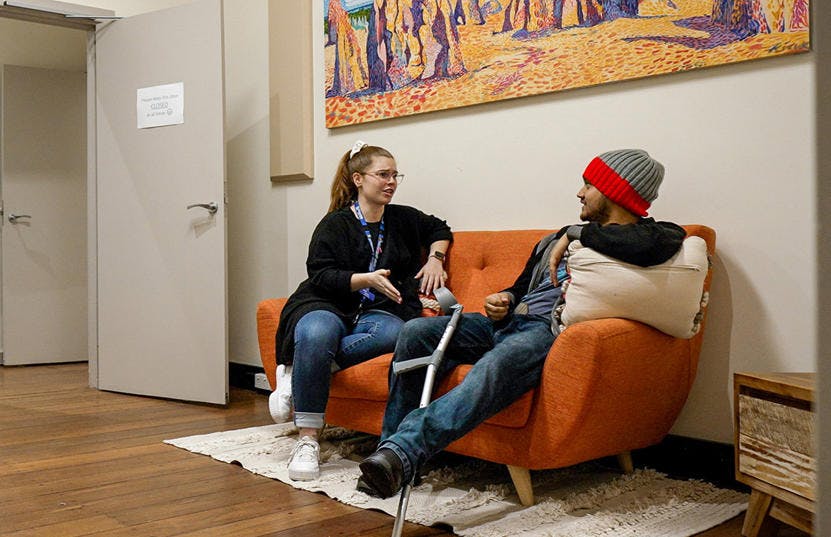 A photo of a Vinnies WA volunteer and a man sitting in an orange couch facing each other and talking. The volunteer is wearing a lanyard, while the man has a crutch leaning against his leg.