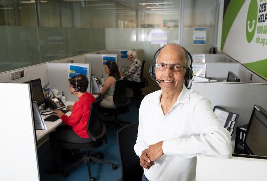 A photo of a man standing in an office landscape. The man is wearing a caller headset and leaning against his office cubicle while smiling at the camera. In the background sits his colleagues in their own cubicles answering calls.
