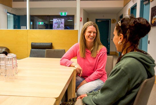 A photo of a Vinnies WA volunteer and a woman sitting down at a table at Tom Fisher house. The volunteer is smiling at the woman, who has her back towards the camera. 