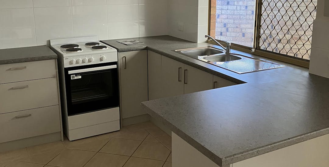A photo of a kitchen showing the countertop, a sink, a stove and an oven. The drawers and cupboards are beige with silver handles, while the sink is silver. There is a window above the sink. 