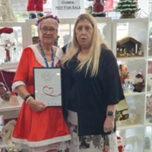 A photo of 2 women, one dressed in a Christmas outfit, standing beside each other facing the camera. One of them is holding a framed acknowledgement.