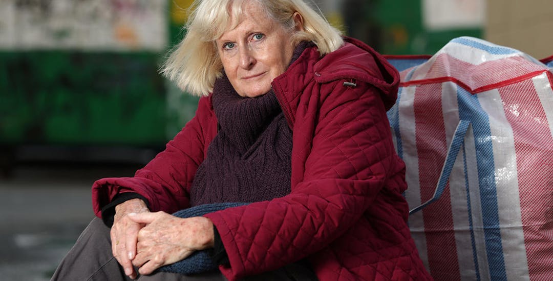 A photo of an elderly woman sitting outside on the pavement with a large bag behind her. She is wearing a red quilted jacket and a black scarf and is facing the camera with her arms around her knees. 
