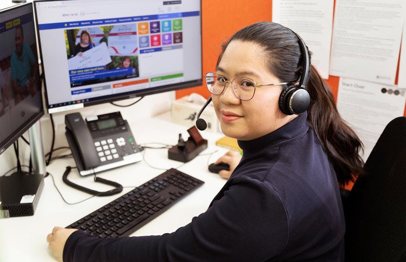 A photo of a Vinnies WA volunteer in her cubicle at the callcentre. She is sitting down by her computer while smiling and facing the camera.