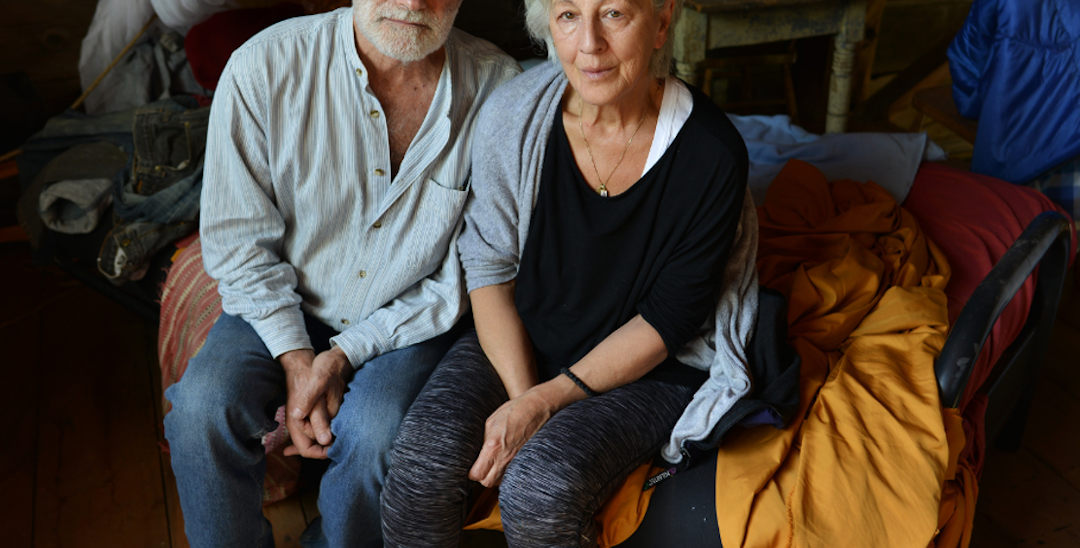 A photo of a man and woman sitting next to each other on a bench with blankets and sheets around them, in casual clothes. They are looking up to the camera.