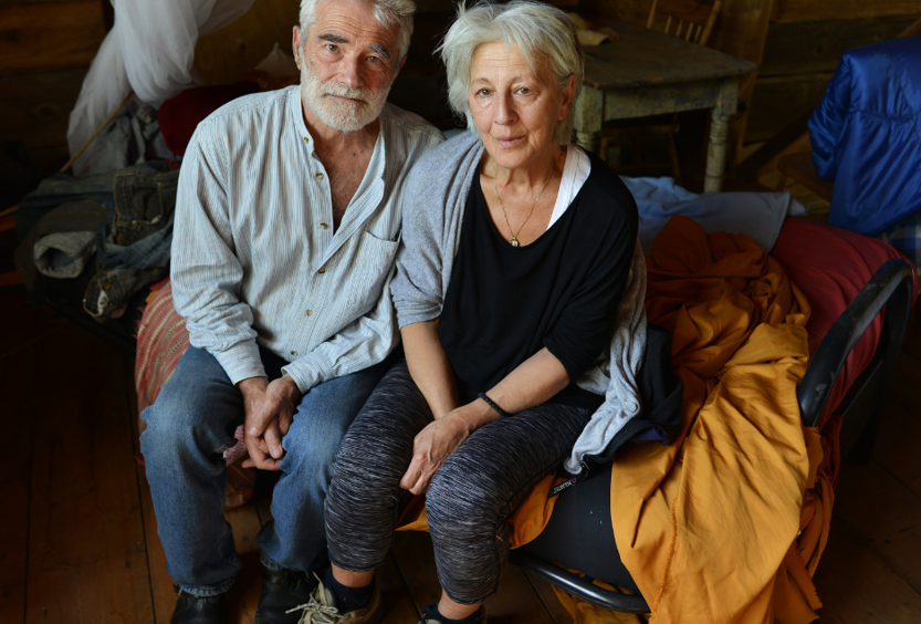 A photo of a man and woman sitting next to each other on a bench with blankets and sheets around them, in casual clothes. They are looking up to the camera.