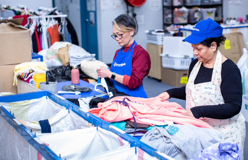 A photo of 2 women at a table sorting clothes and linen. One is wearing a Vinnies cap and the other a Vinies apron.