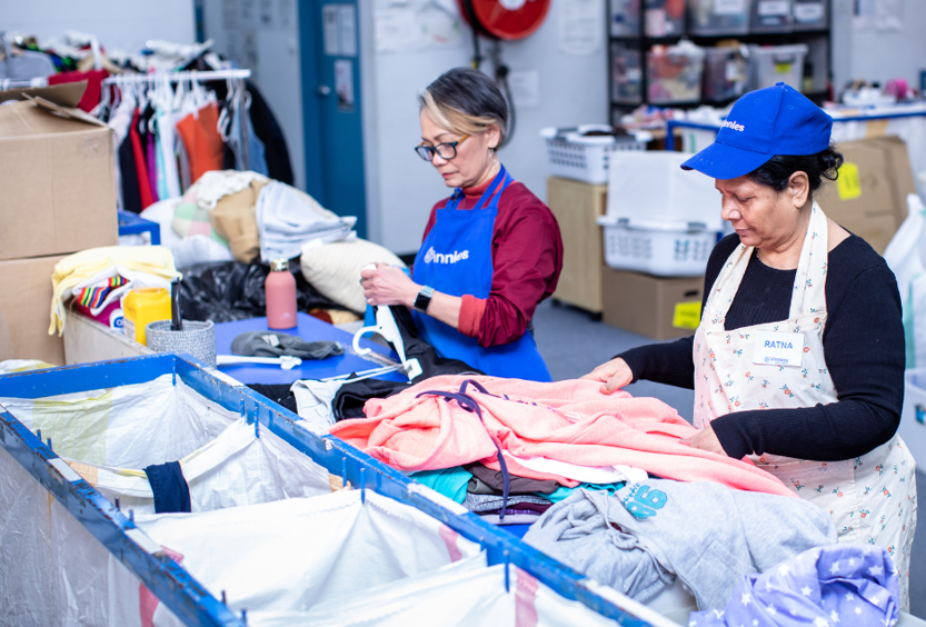 A photo of 2 women at a table sorting clothes and linen. One is wearing a Vinnies cap and the other a Vinies apron.