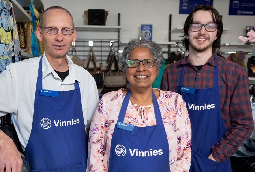 A photo of 3 Vinnies WA Volunteers wearing blue Vinnies aprons. They are at standing inside a Vinnies Shop and some second hand items are displayed in the background. The volunteers are centre of the photo smiling at the camera. 