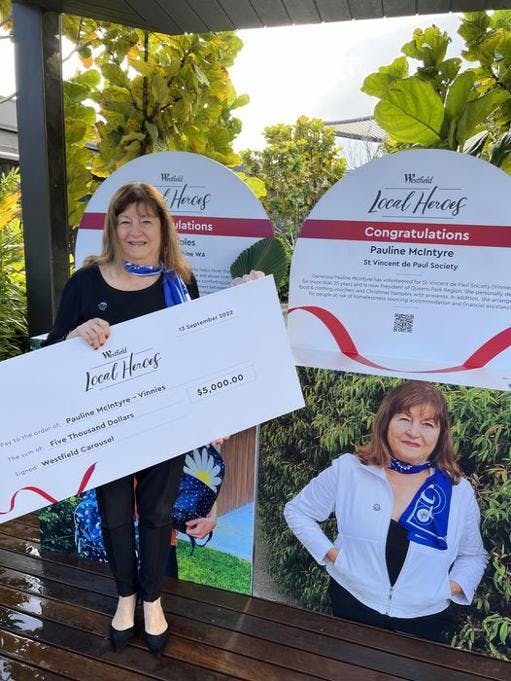 A photo of Vinnies member Pauline McIntyre standing in front of signs congratulating her on her win for the Local Heroes program. She is holding a large check for $5,000.