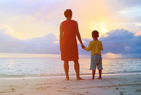 Photo of a woman and a child holding hands on a beach. They are facing away from the camera and looking at a sunset.