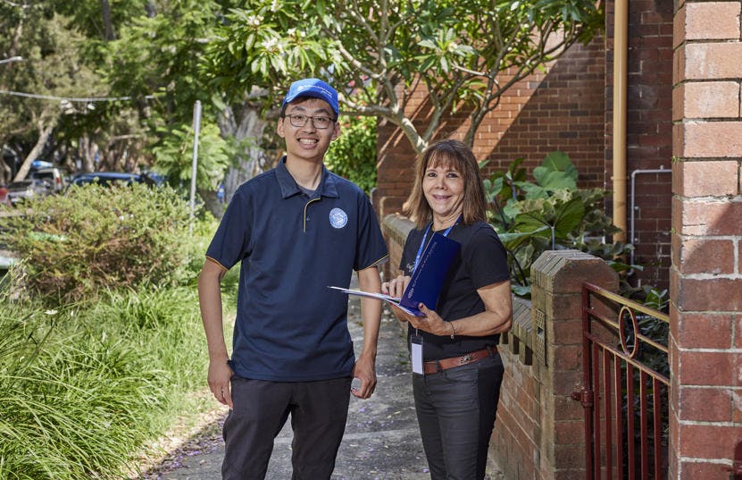 A photo of 2 Vinnies WA volunteers standing outside together facing the camera and smiling. One of them is wearing a lanyard and the other is wearing a blue cap with the St Vincent de Paul Society logo.