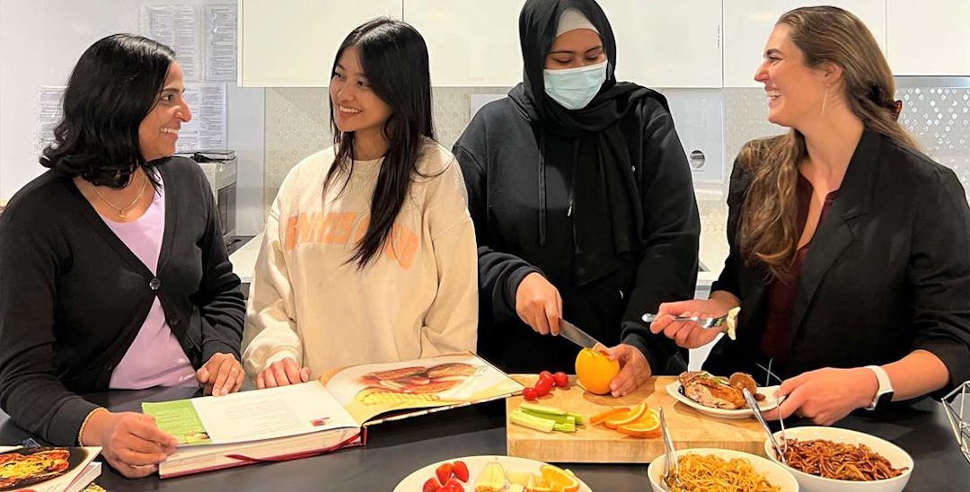 A photo of Vinnies WA staff in the headquarter kitchen. The 4 women are standing behind a kitchen isle with bowls of food on it and a cutting board. The women are smiling at each other while preparing a healthy meal.
