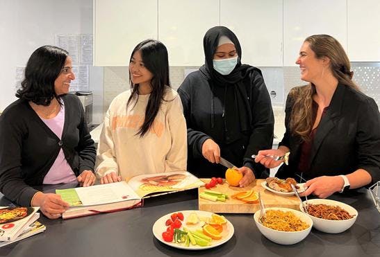 A photo of Vinnies WA staff in the headquarter kitchen. The 4 women are standing behind a kitchen isle with bowls of food on it and a cutting board. The women are smiling at each other while preparing a healthy meal.